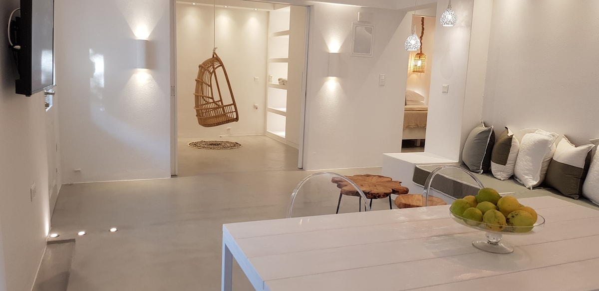 An open-plan living space is depicted, featuring a modern dining table with transparent chairs. To one side, a cozy seating area is arranged with plush cushions. Soft lighting accents the white walls, complemented by a decorative hanging chair in the background.