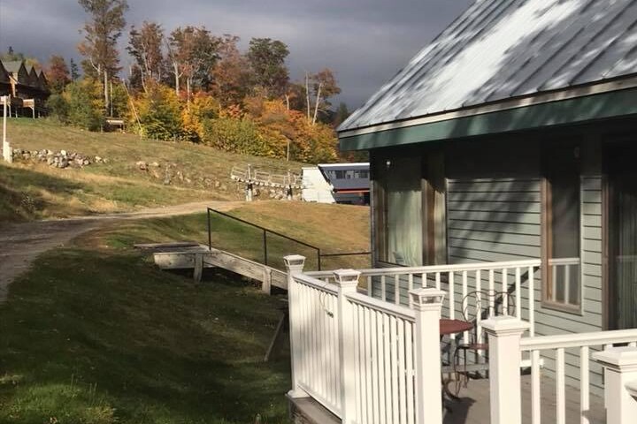 A gentle slope leading away from a building is seen, with vibrant autumn foliage in shades of orange and yellow. A small wooden bridge crosses a path, providing access to the grassy area. In the distance, a shuttle bus is parked near the base of a ski hill.
