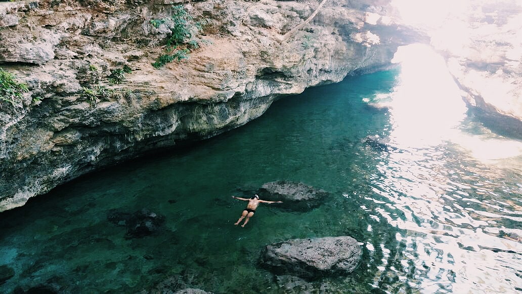 A serene natural pool is depicted, featuring clear turquoise water and rocky formations. Sunlight filters through an opening above, creating a calming environment. A person is seen floating serenely on the surface, enhancing the tranquil setting surrounded by dramatic cliffs.