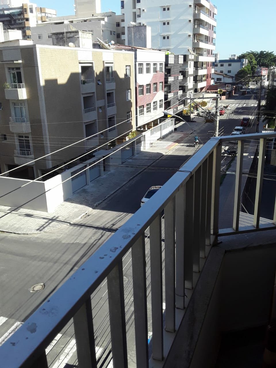 A view from a balcony reveals a city street lined with buildings, with light traffic visible below. A mixture of residential and commercial structures create a vibrant atmosphere. Bright sunlight illuminates the scene, highlighting the clear blue sky above.