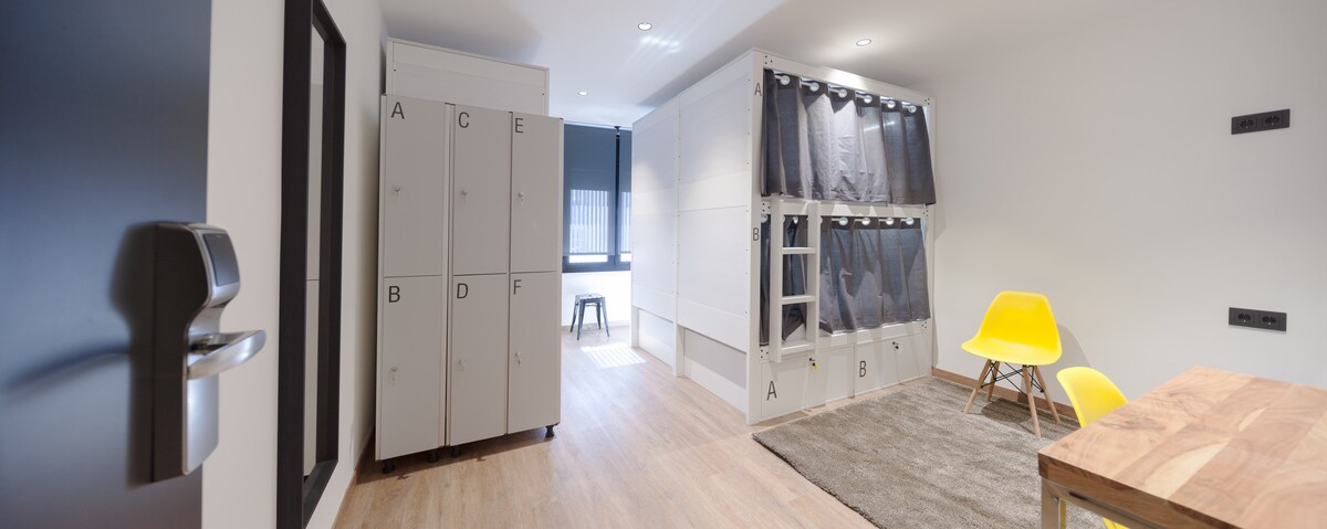 A modern shared room is displayed, featuring organized lockers labeled A to F. Bunk beds with privacy curtains are positioned along the wall. A vibrant yellow chair complements a wooden dining table on a soft area rug, while natural light enters from a window.