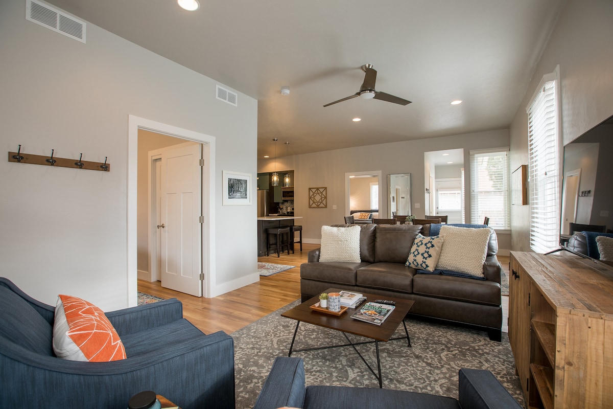 A welcoming living area is presented with a comfortable brown sofa and two accent chairs. A coffee table is centered on a patterned rug, while large windows allow natural light to brighten the space. The open layout connects to a dining area visible in the background.