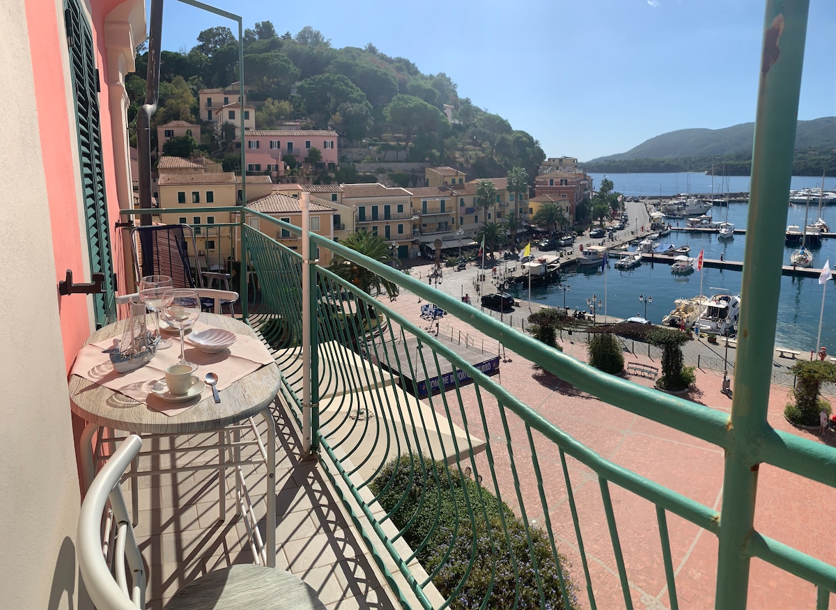 A balcony offers an unobstructed view of the main square and the harbor of Porto Azzurro. Two chairs and a small table are positioned for outdoor dining, with the vibrant colors of nearby buildings and boats enhancing the coastal scenery.