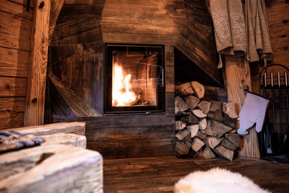 A modern fireplace is centered within a rustic wooden setting, surrounded by stacked firewood and cozy blankets hanging nearby. The warm glow of the flames offers a comforting ambiance, enhancing the inviting atmosphere of the space.