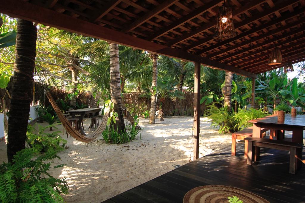 A shaded outdoor area is seen with a wooden table and benches surrounded by lush tropical greenery. A hammock is suspended between palm trees, offering a relaxing space. Soft, sandy ground is visible, contributing to the tranquil nature of the garden.