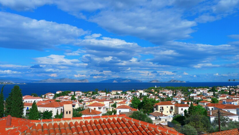 A panoramic view showcases the coastal village of Dapia, featuring terracotta-roofed buildings nestled among lush greenery. The serene blue waters of the Mediterranean Sea stretch into the horizon, under a vibrant sky with scattered fluffy clouds.
