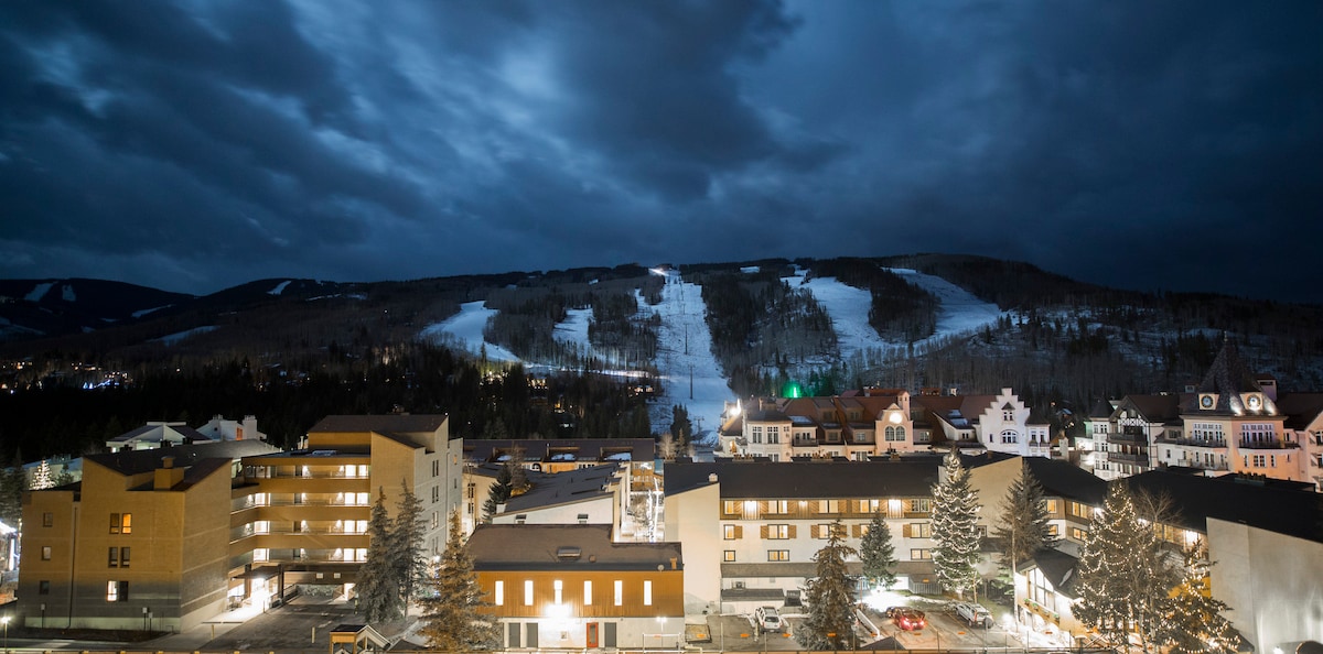 A panoramic view captures a winter evening scene, showcasing a lit-up community with familiar buildings. Snow-covered slopes ascend in the background, framed by a subtle mountain skyline beneath a cloudy sky.