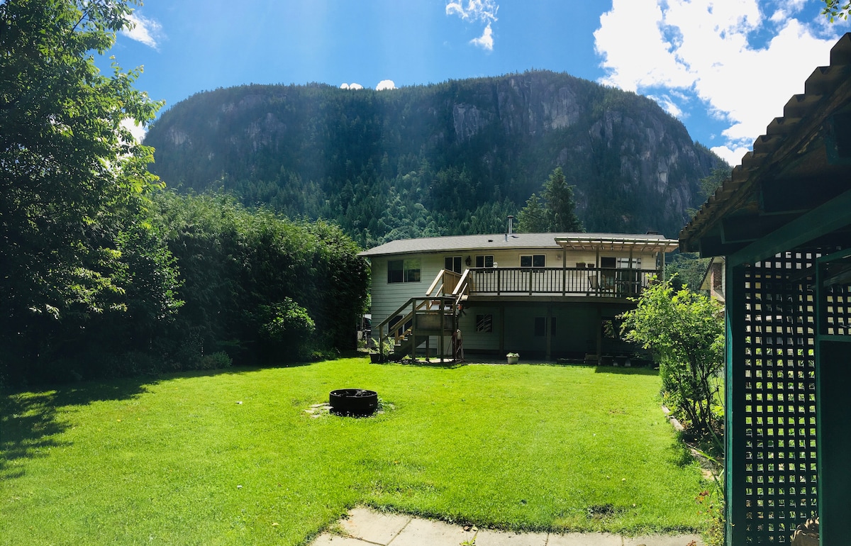 The expansive backyard offers a well-maintained lawn surrounded by lush greenery. A fire-pit sits at the center, with the inviting deck of the ground-level suite visible in the background. Majestic mountain views complete the serene outdoor setting.