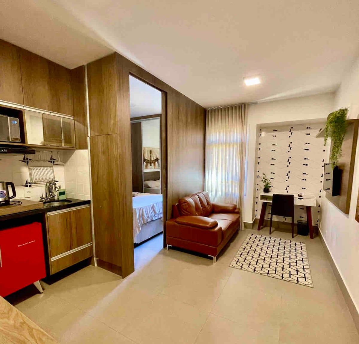 The studio layout features a kitchenette with modern appliances, including a microwave and coffee maker. A cozy seating area is highlighted by a brown sofa. Natural light filters through the large window, illuminating the workspace with a desk and chair, while a partition separates the sleeping area.