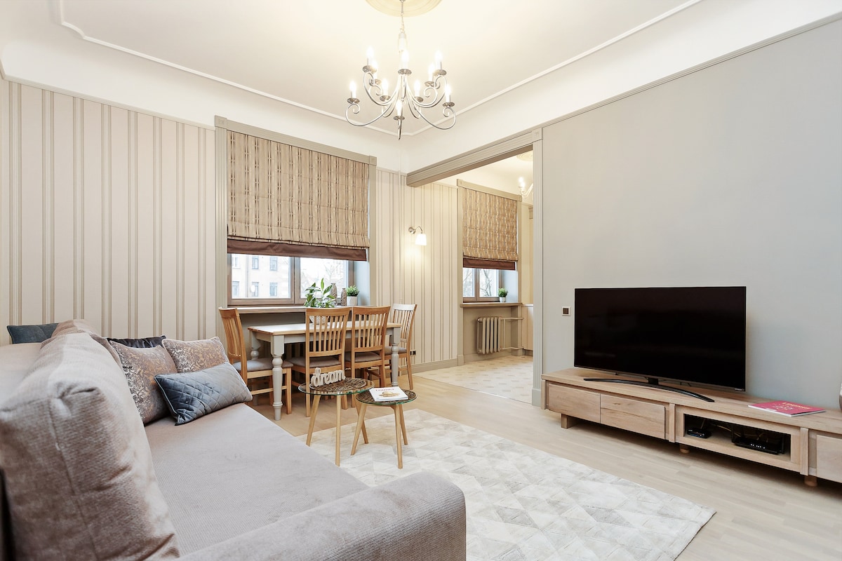 A light-filled living area features a comfortable sofa adorned with decorative pillows. A dining table with chairs is positioned to one side, and a flat-screen television is mounted on a wooden stand. Soft wood accents complement the subtly patterned walls.