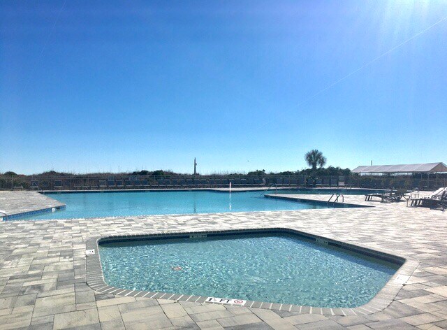 An outdoor swimming pool area is visible, featuring a spacious main pool accompanied by a smaller hot tub. Sunlight reflects off the water surface, and several loungers are positioned nearby on a tiled deck. Clear blue skies are seen overhead, enhancing the open atmosphere.