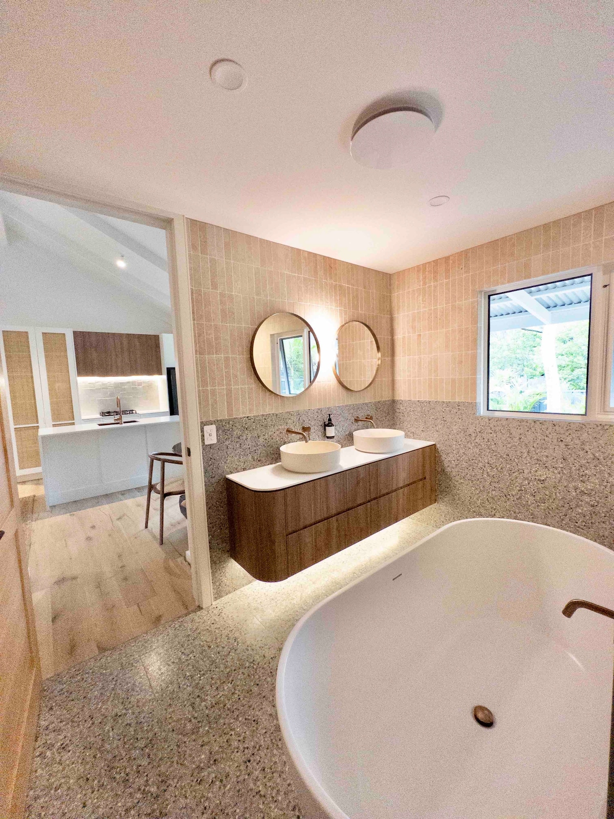 A luxurious bathroom is highlighted by dual circular mirrors above a floating vanity with double basins. A soaking tub is positioned in front of a window, surrounded by natural stone tiles in soft hues, creating a tranquil space filled with natural light.