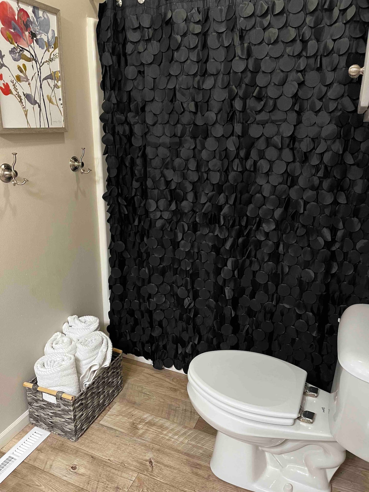 A bathroom setting features a white toilet beside a light-colored wall. A decorative curtain with circular patterns partially conceals the shower area. A woven basket holds neatly stacked white towels, while two hooks on the wall provide additional hanging space.