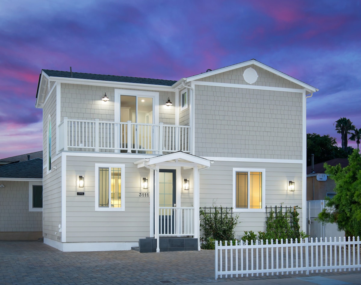 The two-story house features a light-colored exterior and distinctive shingle siding. A front porch is accessed by steps, with a door framed by two glass panels. A white picket fence borders the well-maintained yard, complemented by evening lighting against a colorful sky.