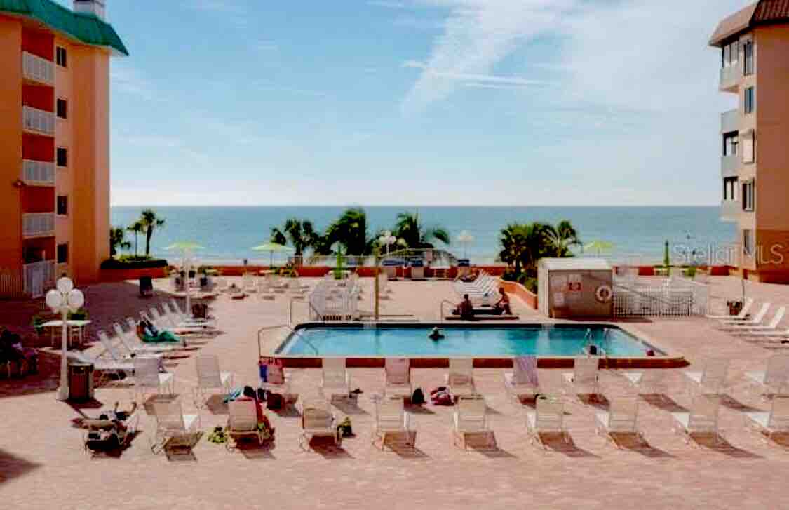 The pool area is surrounded by lounge chairs, with palm trees and ocean views in the background. The inviting swimming pool is centrally located, and sun umbrellas provide shade. A hot tub is positioned nearby, contributing to a relaxing outdoor atmosphere.