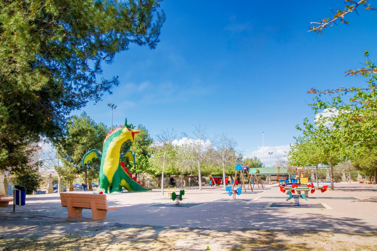 A vibrant park is featured with colorful playground equipment, including swings and slides. A large, whimsical dinosaur sculpture is present among the lush green trees. Benches for seating are positioned throughout the area, offering places to relax and enjoy the outdoor space.