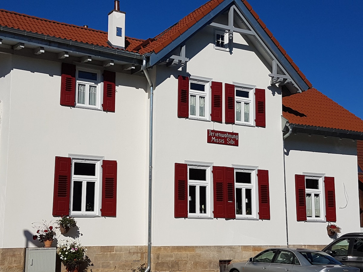 The exterior of the building features a white facade complemented by red shutters and a tiled roof. The entrance is marked by a sign that reads 'Ferienwohnung Missis Söh'. Flower pots are placed by the windows, contributing to the overall charm of the property.