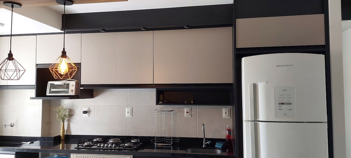 A modern kitchen featuring sleek cabinetry in neutral tones and a spacious countertop. Two pendant lights provide illumination, while a gas stove and an oven microwave combo are visible. A refrigerator stands prominently, and thoughtful organizational tools can be noticed on the countertop.