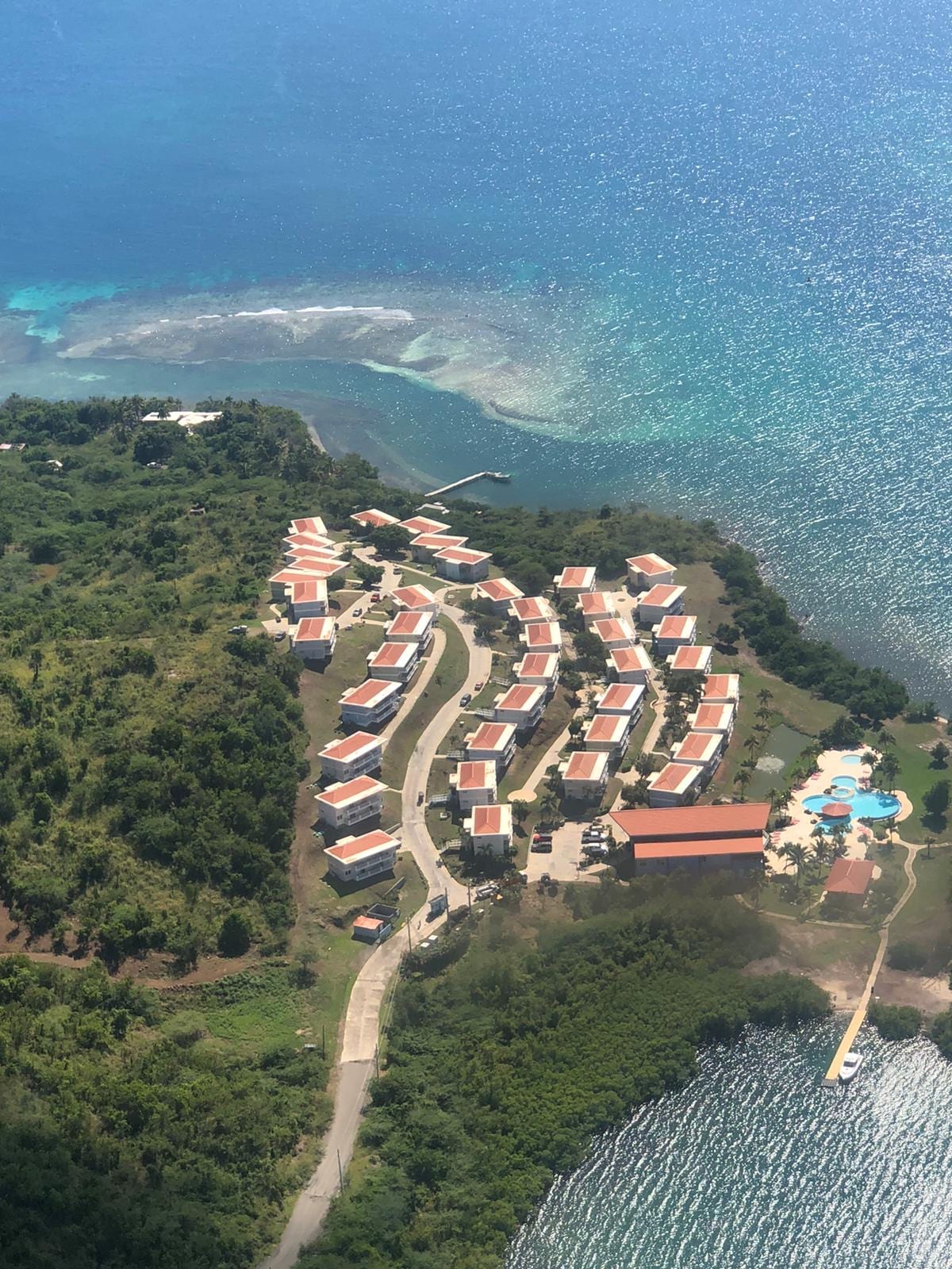An aerial view captures a collection of buildings with red roofs arranged in a curved layout near a shoreline. The azure water surrounds the area, with a pool visible in the foreground. Lush greenery contrasts with the vibrant colors of the architecture.