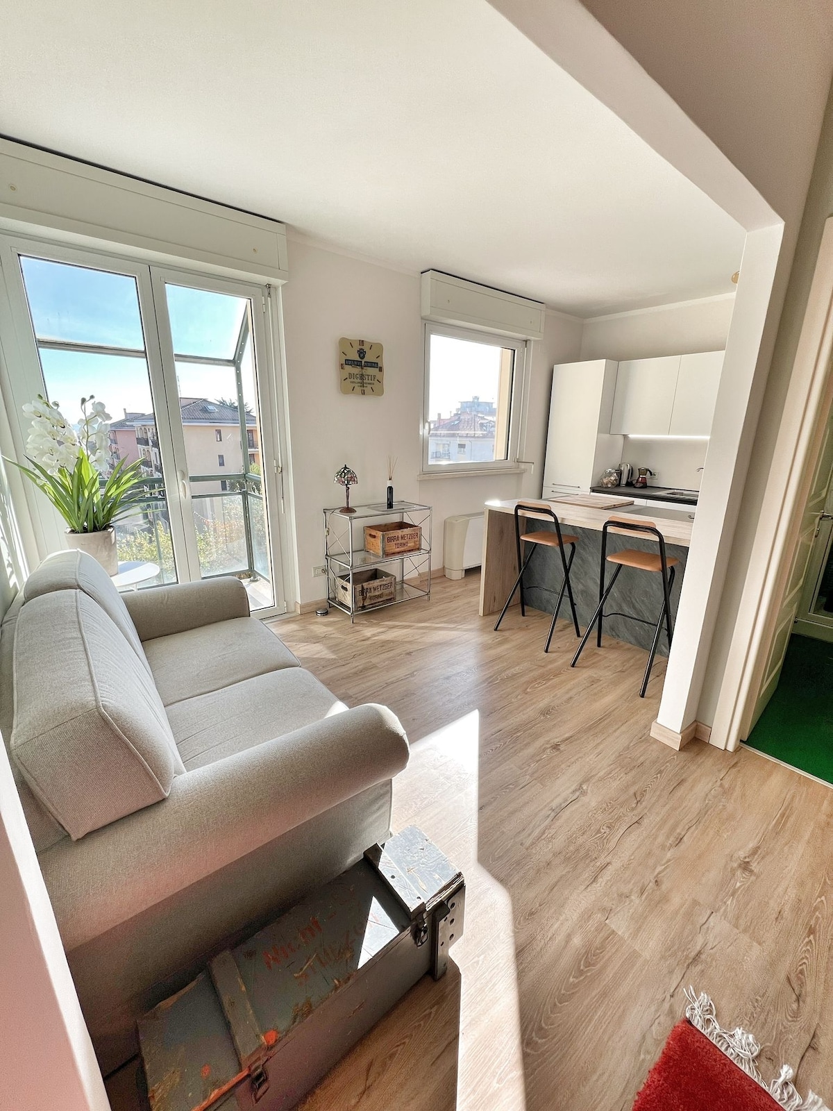 A bright living area features large glass doors opening to a balcony, allowing natural light to fill the space. A comfortable couch is placed next to a small dining table and chairs, with a kitchenette visible in the background. Decorative elements add a touch of warmth.