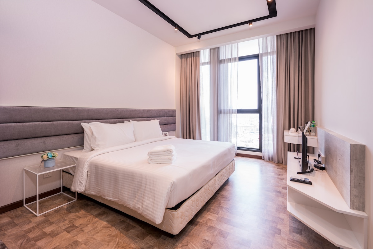 A spacious bedroom features a king size bed with neatly arranged white linens and towels. Large windows allow natural light to fill the room, offering views of the surrounding area. Light wooden flooring complements the contemporary furnishings, creating a calm and clean environment.