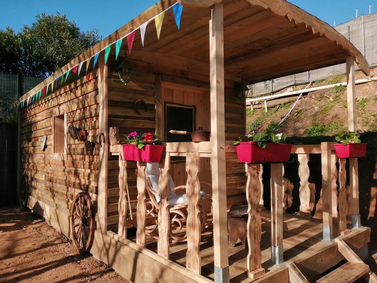 A rustic wooden cabin features a welcoming porch adorned with flower boxes and colorful bunting. The structure showcases natural wood finishes, blending harmoniously with the surrounding landscape. A decorative wagon wheel lends a charming touch to the exterior, enhancing the cabin's unique character.