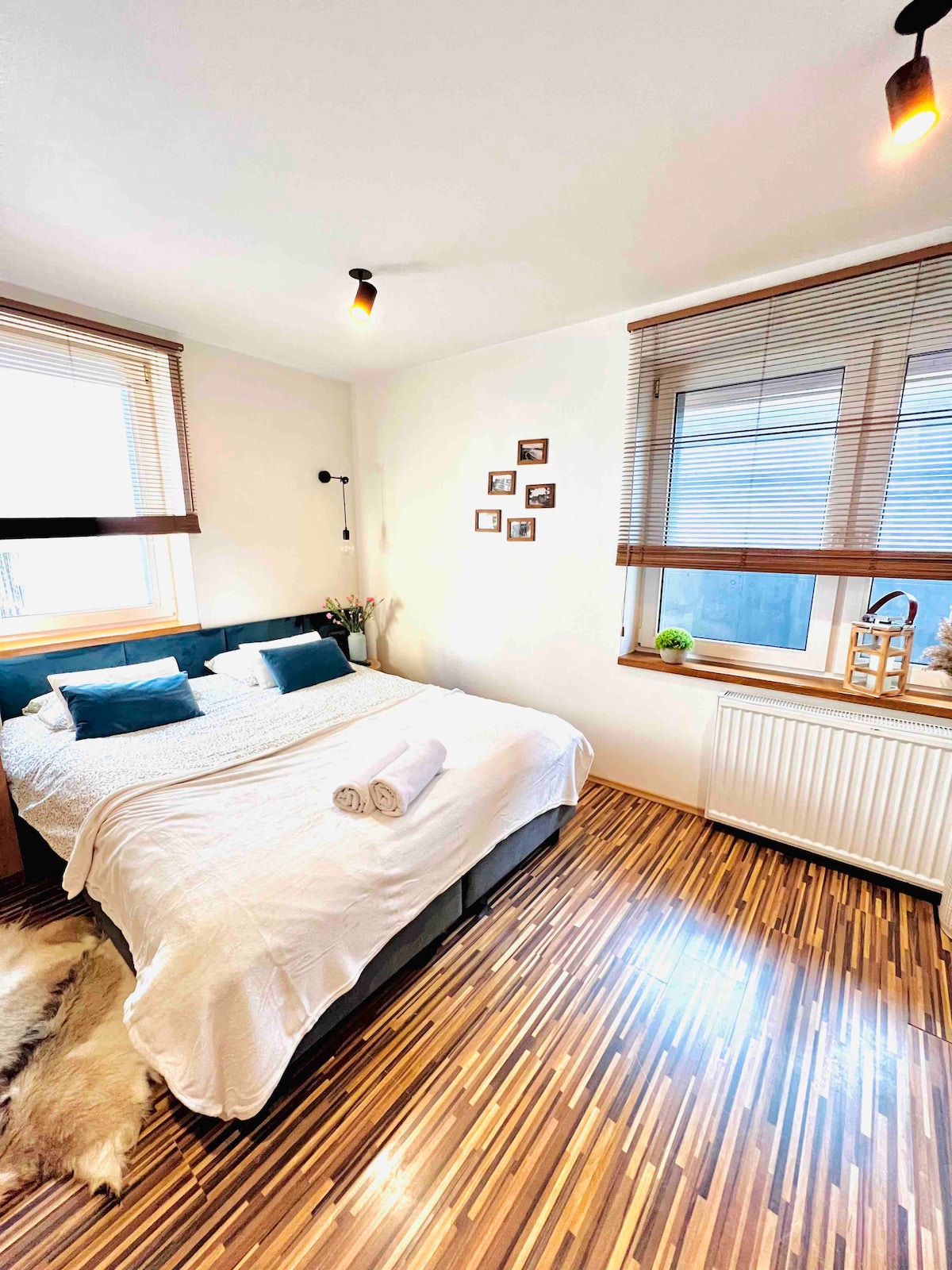 A spacious studio room is featured, showcasing a comfortable bed with soft linens and decorative pillows. Natural light enters through a window adorned with blinds. A smooth wooden floor enhances the cozy ambiance, while framed photographs add subtle decor to the wall.