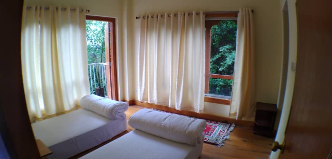A well-lit room features two spacious beds, with rolled towels placed at the foot of each bed. Large windows are adorned with sheer curtains, allowing natural light to illuminate the space. A small rug is placed on the wooden floor, adding warmth to the area.