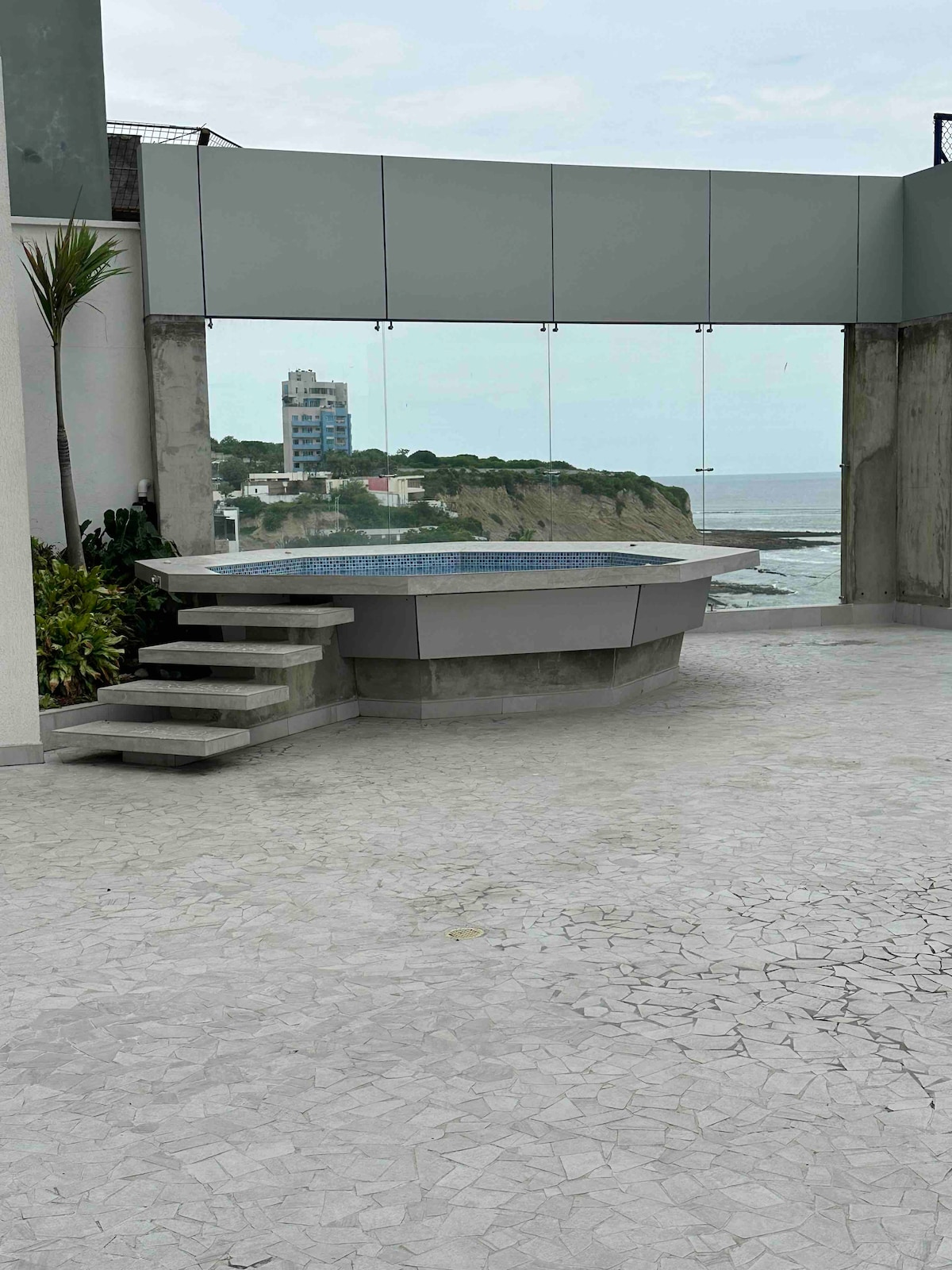 A circular hot tub is positioned on a spacious terrace, bordered by lush plants. Smooth steps lead to the tub, while expansive glass panels provide unobstructed ocean views. The surrounding area features a textured stone floor, enhancing the serene coastal atmosphere.