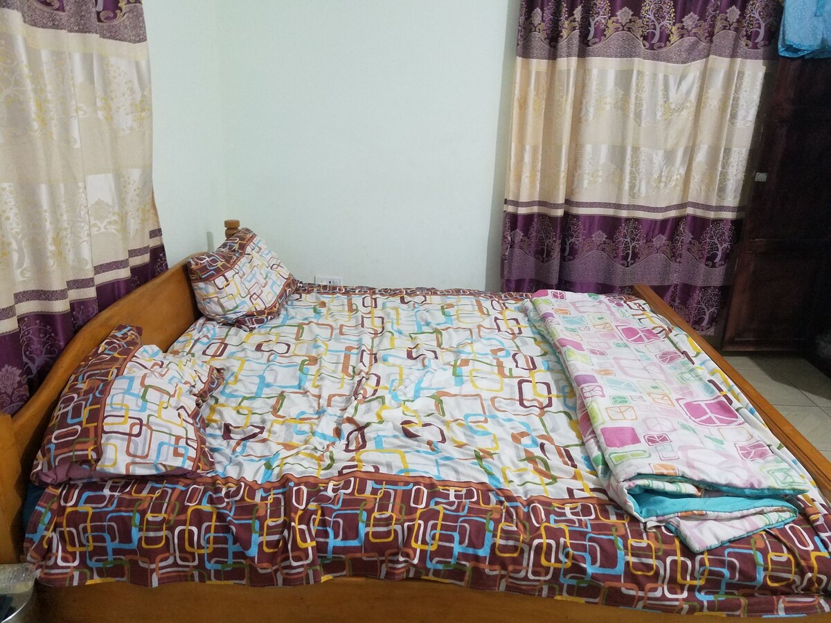 A spacious bed is presented with a colorful patterned bedspread and matching pillows. Soft curtains in shades of purple frame the window, providing a gentle ambiance. A neatly folded blanket lies on the bed, complementing the overall decor.