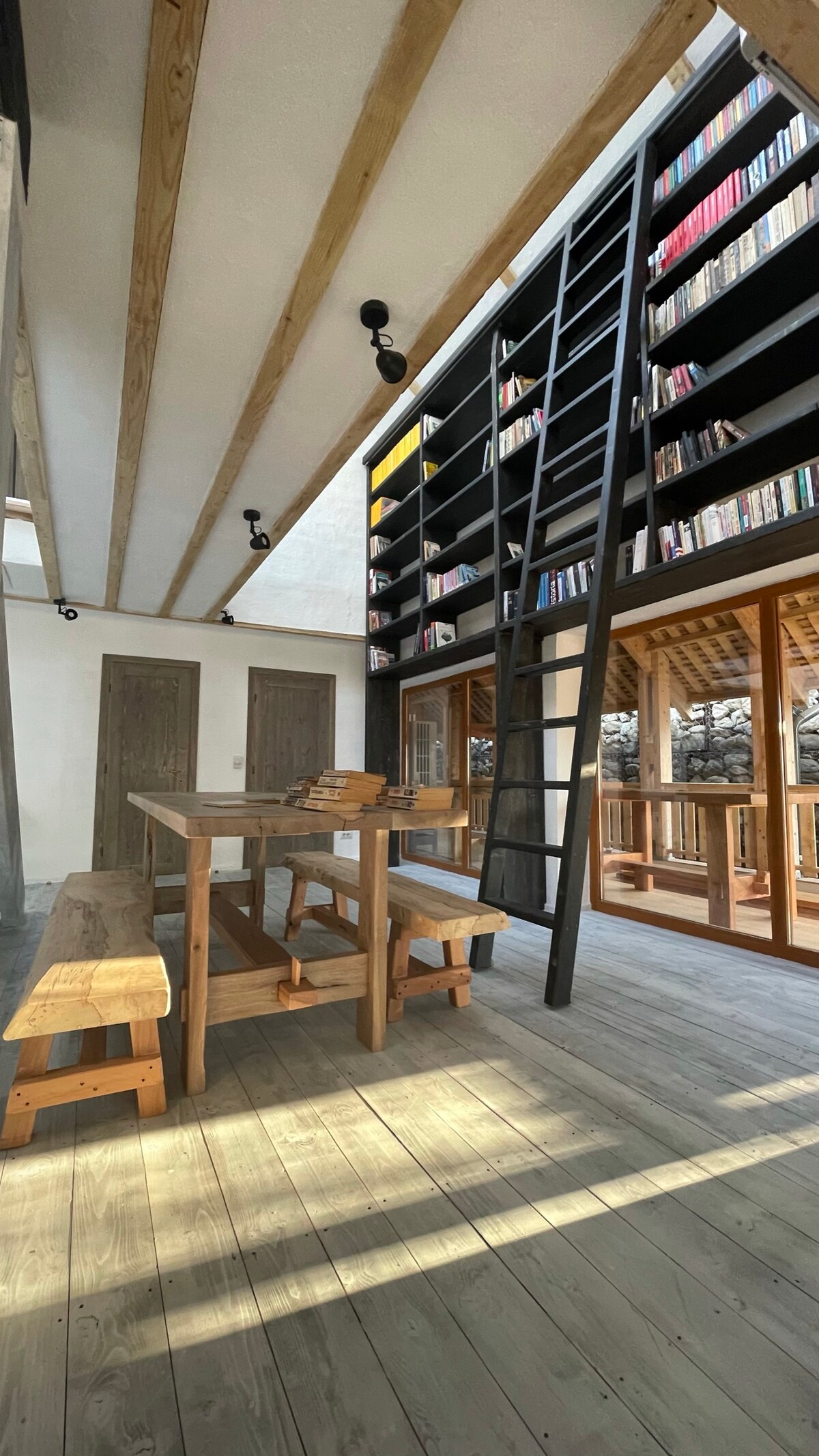 A bright and airy library space is showcased, featuring tall black bookshelves filled with books. A wooden table and benches are positioned centrally on the light-colored wooden floor, while large windows allow natural light to filter in and brighten the area.