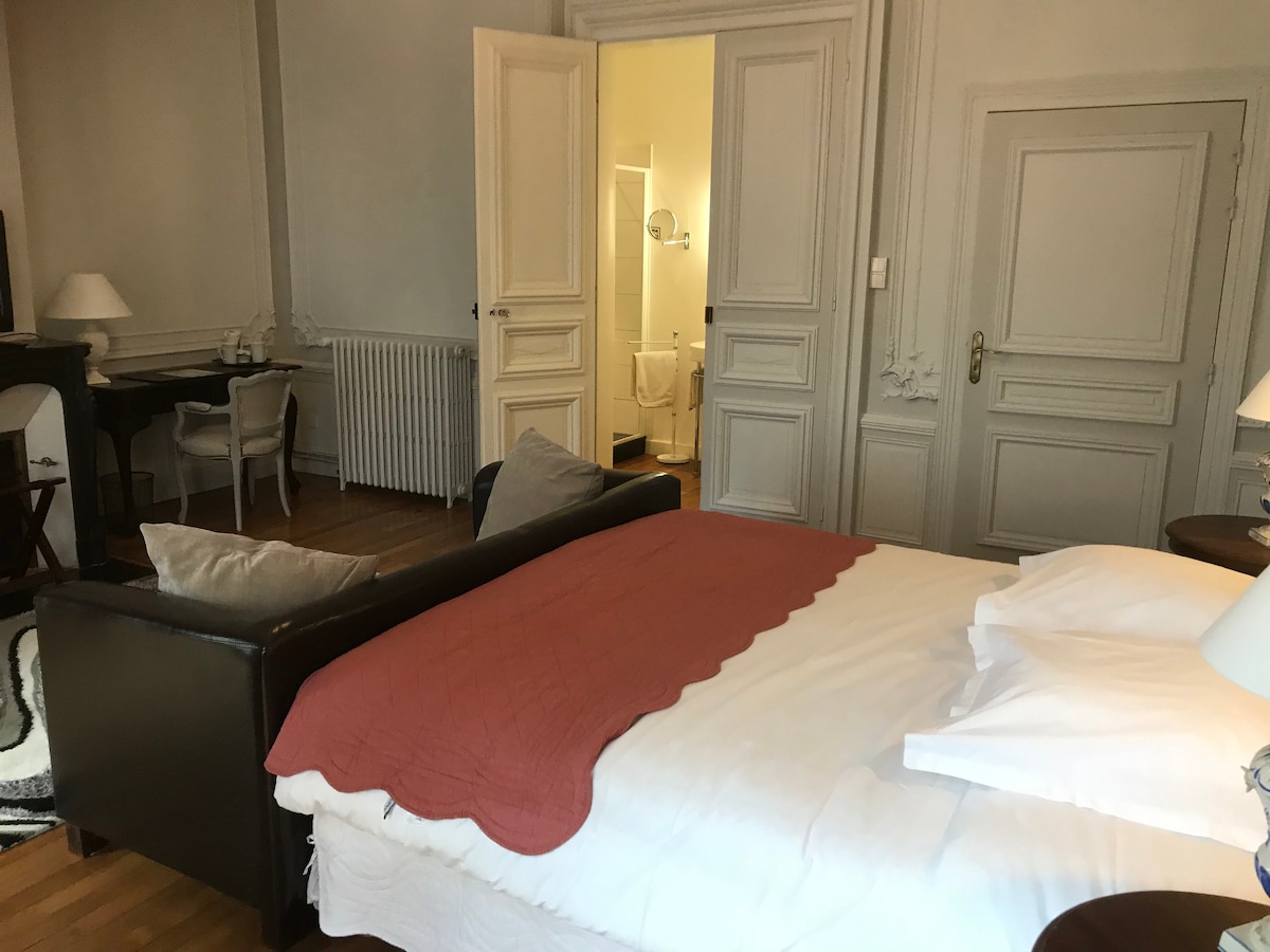 A spacious room is featured, highlighted by a large bed adorned with a coral-colored throw. A comfortable seating area with light-colored pillows is present. A desk and lamp create a functional workspace, while elegant doors lead to an en-suite bathroom, contributing to the room's charm.