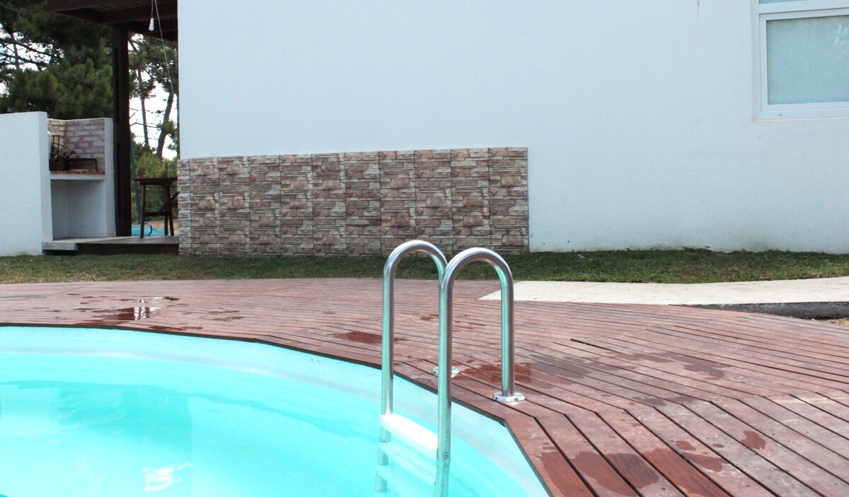 A refreshing outdoor pool is framed by a wooden deck, featuring stainless steel steps leading into the water. A portion of a white building and a stone wall are visible in the background, along with grassy areas surrounding the pool.