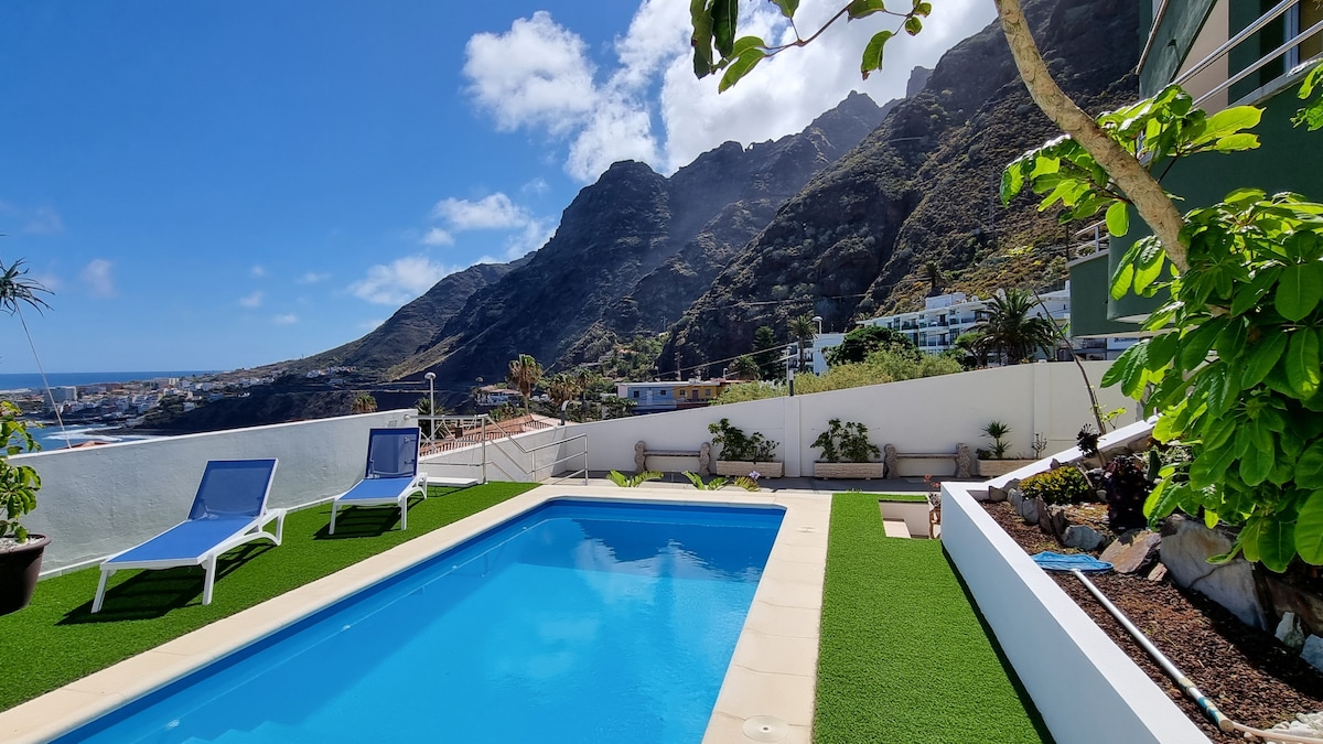 A private pool area is surrounded by lush green grass and features two lounge chairs positioned for relaxation. The clear blue water of the pool reflects the sky, while the backdrop showcases majestic mountains and a view of the coastal landscape.