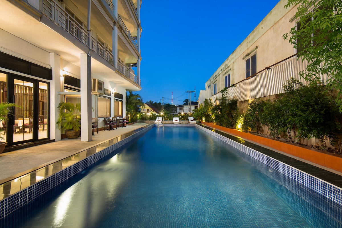 A long outdoor swimming pool is surrounded by well-maintained greenery, with lounge chairs positioned along the water's edge. The area is illuminated softly by ambient lighting, while the building's modern architecture complements the tranquil setting. The evening sky is painted in deep blue hues.