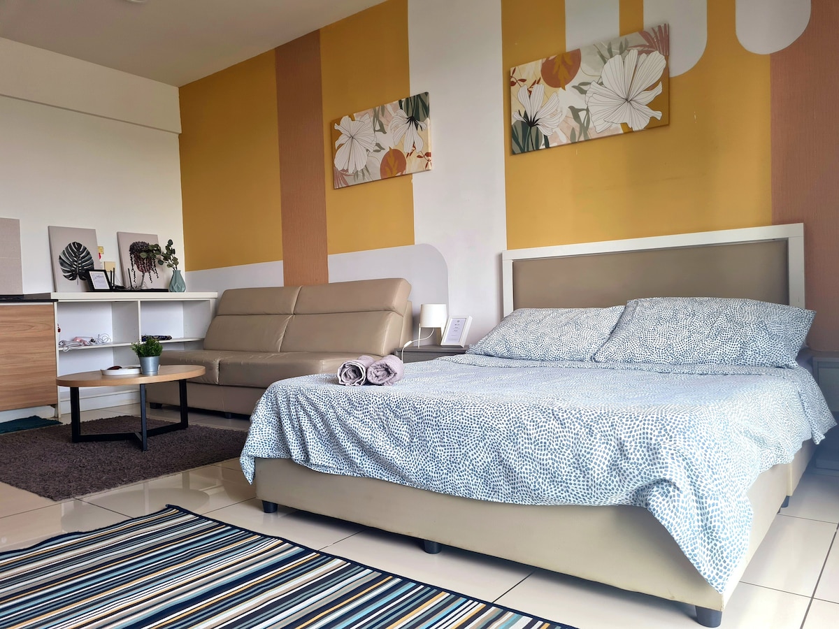 A well-arranged living space features a spacious bed with a patterned coverlet, complemented by a beige sofa. Decorative wall art adds color against a warm yellow backdrop. A round table sits nearby, with fresh towels neatly arranged, and a striped area rug adds texture to the floor.