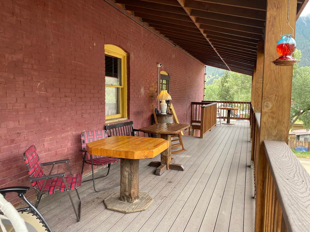 A covered porch features wooden flooring with a rustic table at its center. Two chairs are arranged around the table, complemented by additional seating along the railing. The backdrop showcases a red brick wall, creating a warm and inviting outdoor space.