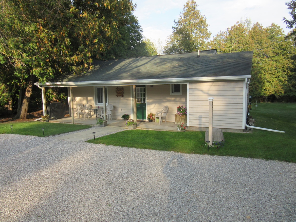 The single-story cottage features a covered front porch with several chairs arranged for seating. A gravel pathway leads to the entrance, surrounded by a well-maintained lawn and trees. A subtle landscape design includes decorative plants and a peaceful outdoor setting.