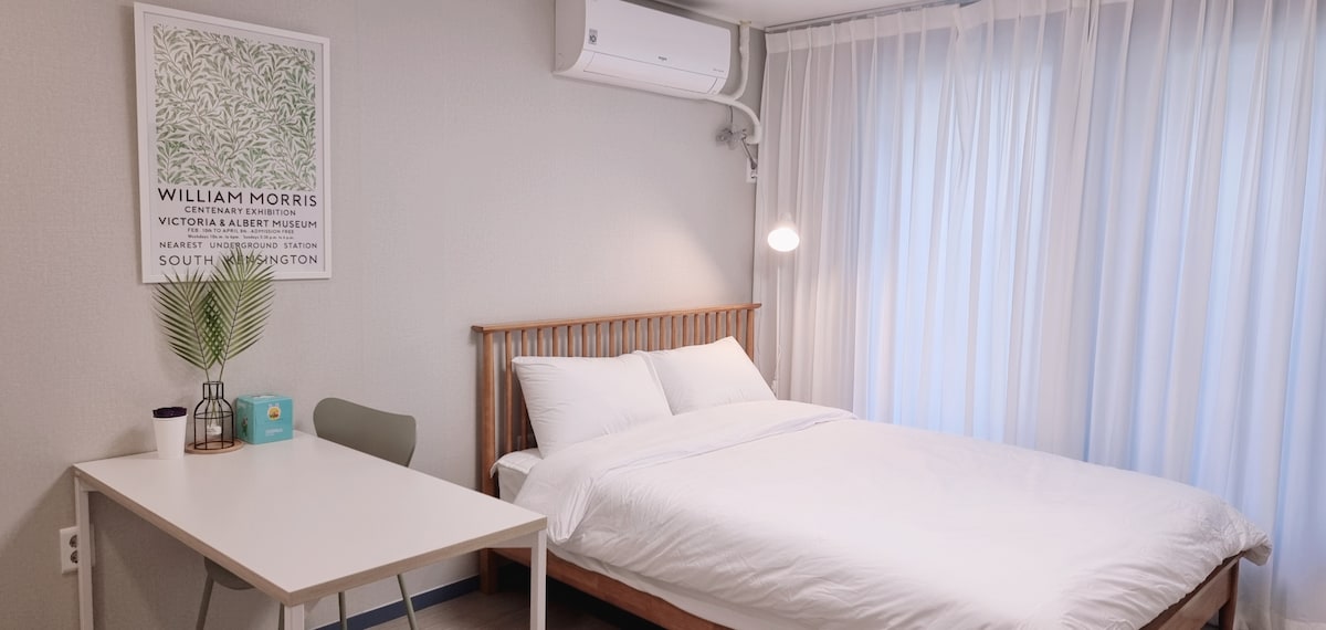 A bright and neatly arranged bedroom is highlighted by a spacious bed with fresh linens. A small desk and chair are positioned nearby. Large window drapes allow natural light to fill the room. A framed artwork adorns the wall, enhancing the serene atmosphere.