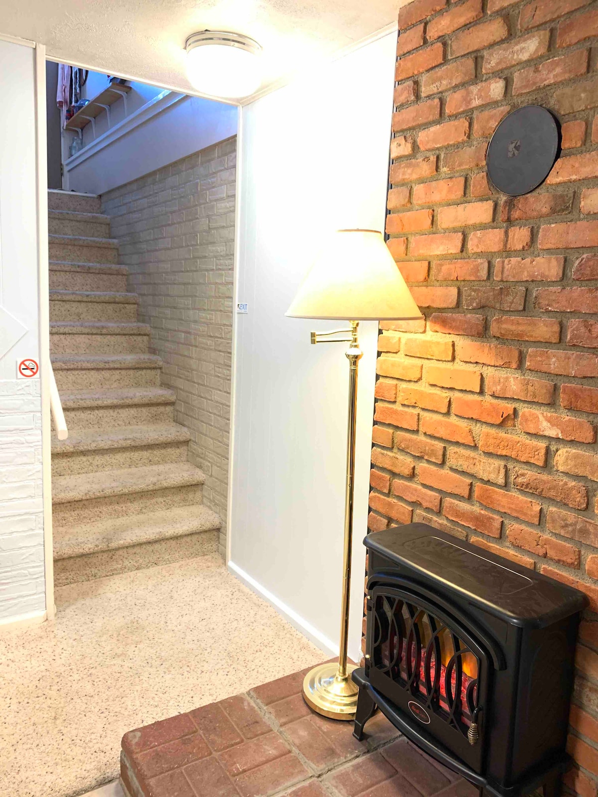 A well-lit hallway is showcased, featuring a staircase leading to the upper level. A small electric fireplace sits beside a floor lamp against a brick wall, providing warmth and illumination. The inviting atmosphere is complemented by neutral-colored walls and a clean, tidy floor.