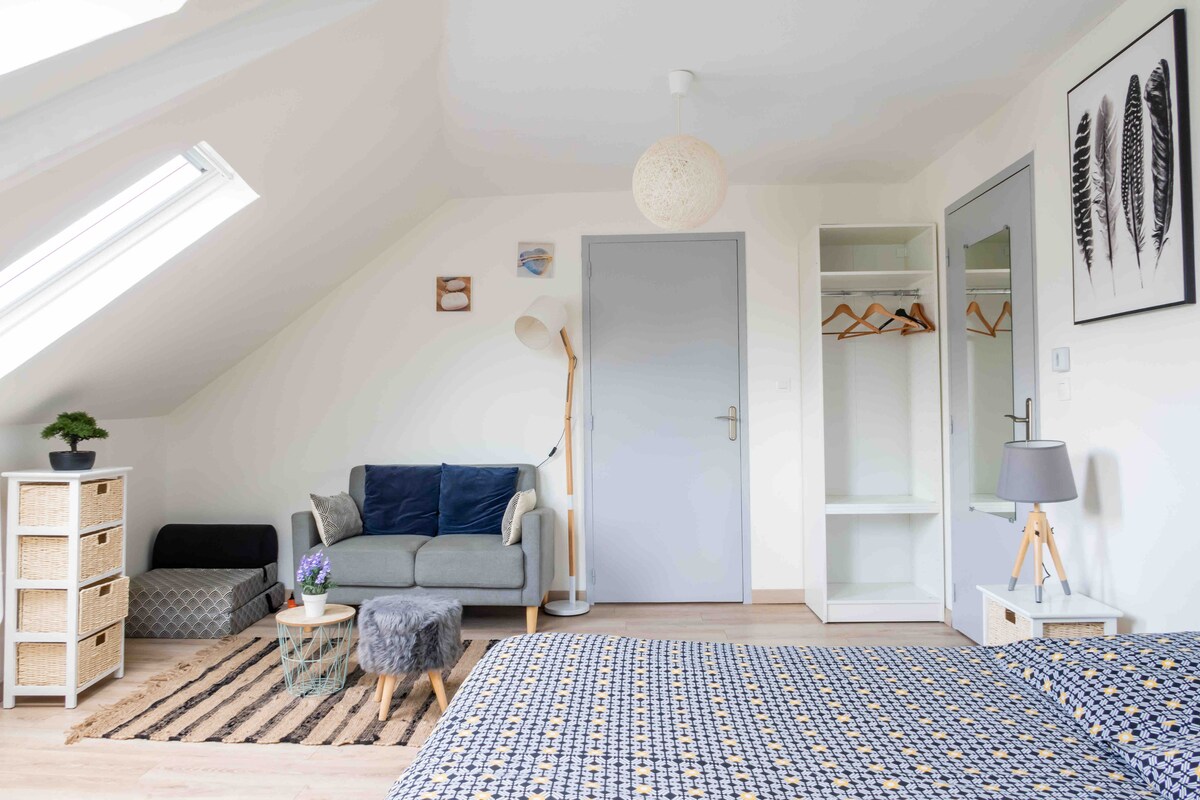 A bright, inviting room is featured, showcasing a spacious bed with a patterned coverlet. A cozy sofa and a stylish lamp are positioned nearby. A closet with hanging space and a dresser add functionality. Natural light filters through a skylight, enhancing the open atmosphere.