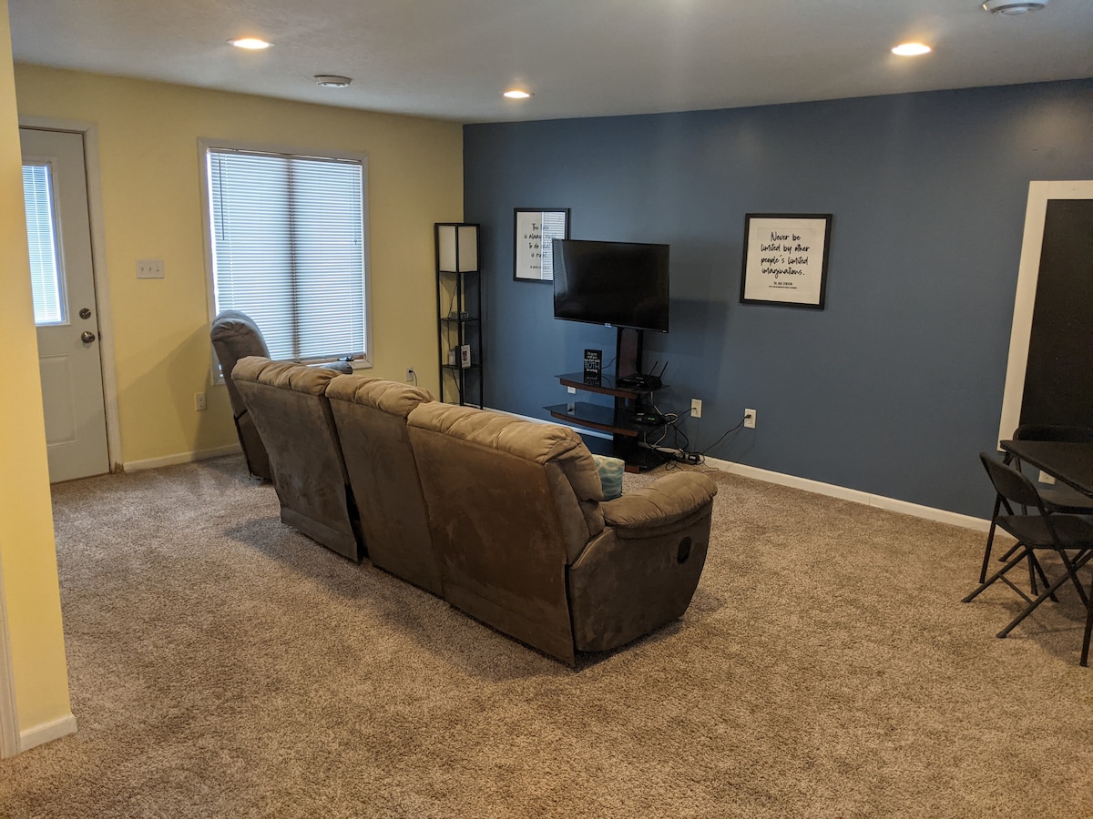 A spacious living room is viewed, featuring a comfortable sectional sofa in a neutral tone. A flat-screen TV is positioned against a blue wall, complemented by decorative artwork. Natural light filters through a window, while a small dining area is visible in the corner.