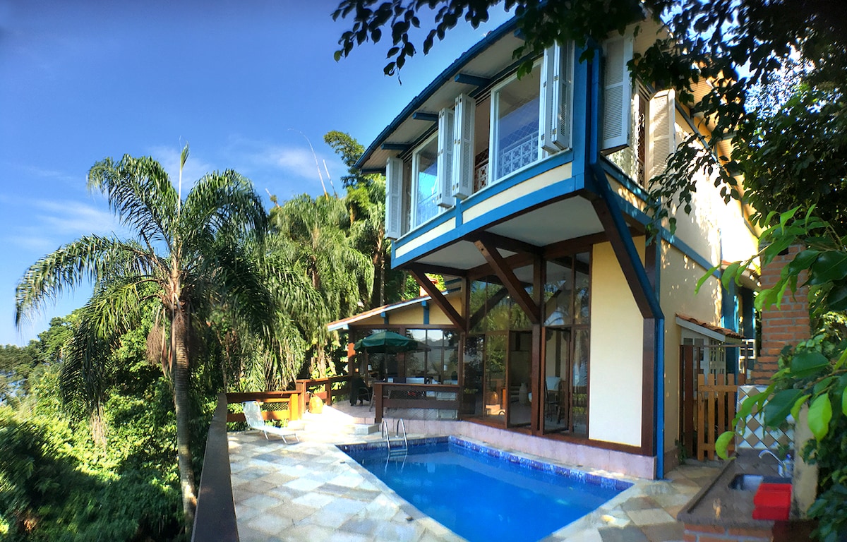 The two-story house is surrounded by lush greenery, featuring a private pool in front. Large glass windows and traditional shutters are present, allowing natural light into the interior. A wooden deck extends from the house, providing a space for relaxation.