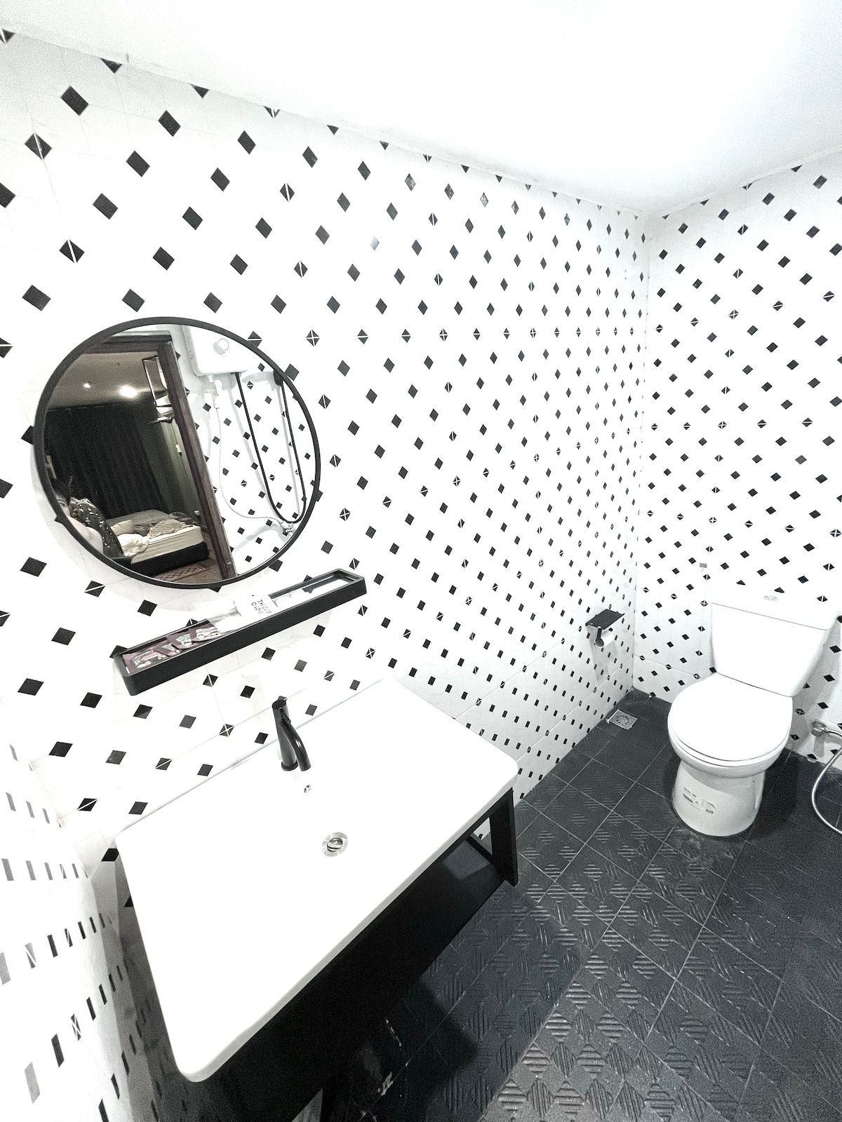 The bathroom features a modern design with black and white diamond-patterned tiles. A round mirror is mounted above a sleek sink, complemented by a minimalist black countertop. A white toilet is positioned against one wall, providing a clean and functional space.