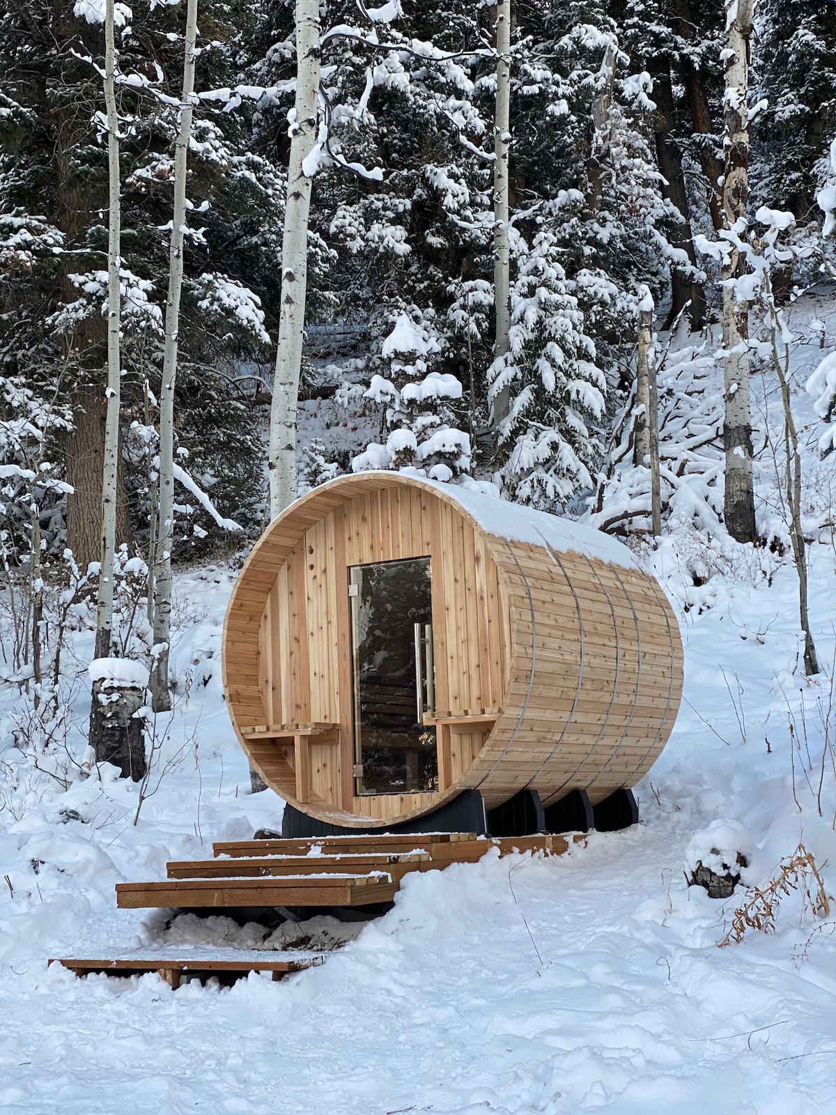 An outdoor barrel sauna is nestled among snow-covered trees, showcasing a wooden structure with a rounded design. Steps lead to the entrance, which is framed by a layer of fresh, white snow, providing a serene winter atmosphere.