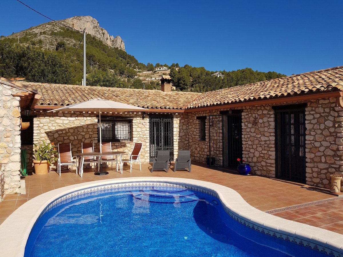 A private courtyard features a clear swimming pool surrounded by terracotta tiles. A shaded dining area with a table and chairs is positioned next to the pool. Sun loungers are arranged for relaxation, with stone walls and a mountainous landscape visible in the background.
