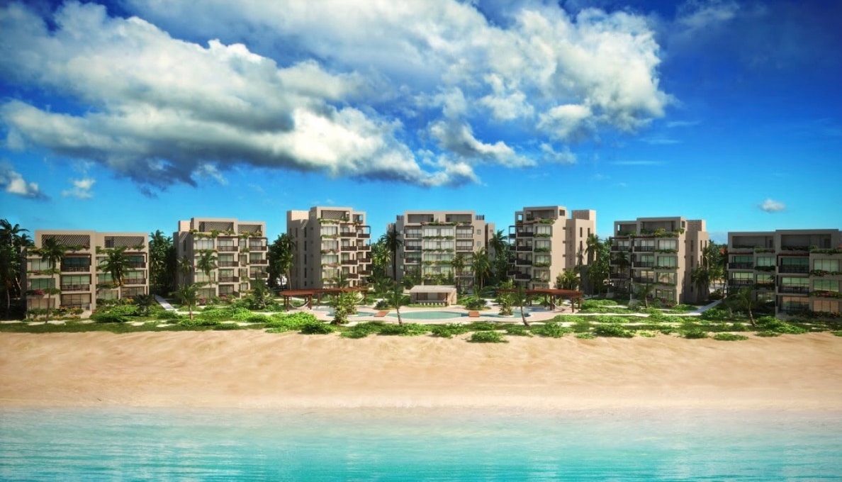 A beachfront view captures a collection of modern buildings surrounded by lush greenery. A central pool area is visible in the foreground, complemented by a sandy beach that leads to clear water. The sky features scattered clouds against a bright blue background.