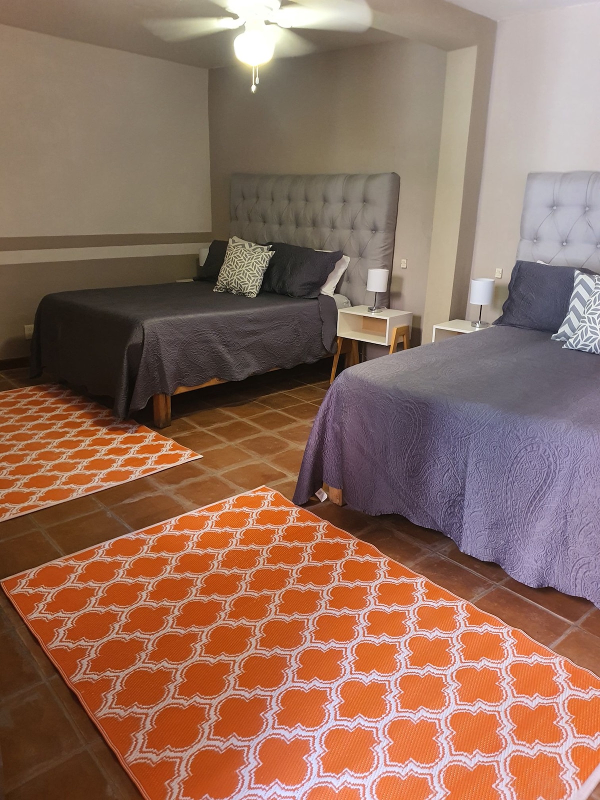 A spacious room contains two stylish beds, each with a tufted headboard and coordinating bedding. Two orange geometric patterned rugs are positioned on the tiled floor, complementing the overall decor. A ceiling fan is visible, providing additional comfort in the space.