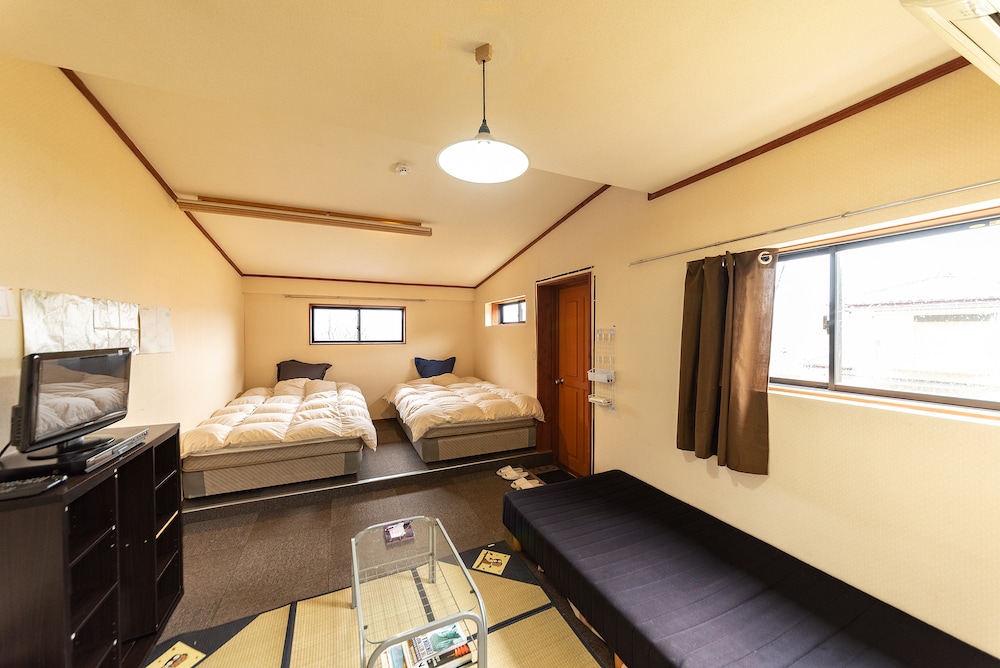 The room features two spacious beds with soft bedding arranged on separate platforms. A low table sits in the center, accompanied by a dark sofa. Natural light enters through two windows, while a TV and storage unit provide additional amenities.