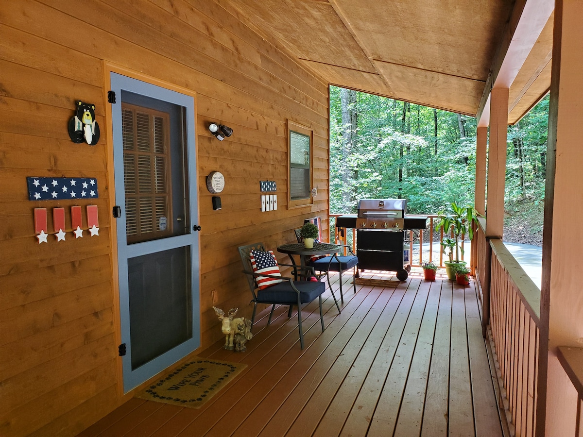 The front porch features a wooden floor and natural wood paneling, accented by outdoor seating arrangements that include two chairs and a small table. A grill is positioned nearby, and decorative elements reflect a subtle patriotic theme, creating an inviting outdoor space surrounded by trees.